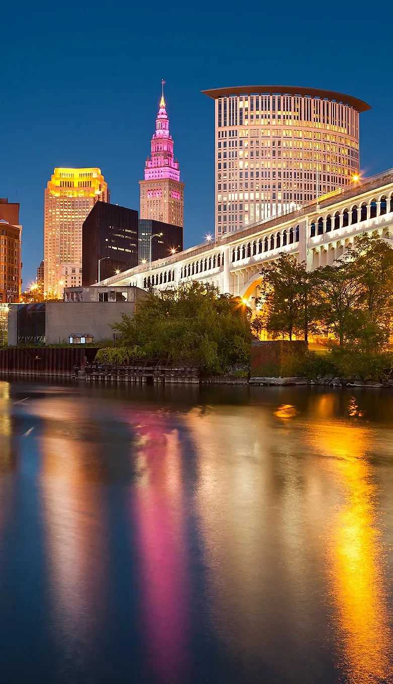 City skyline at dusk with illuminated buildings and bridge reflections in water.